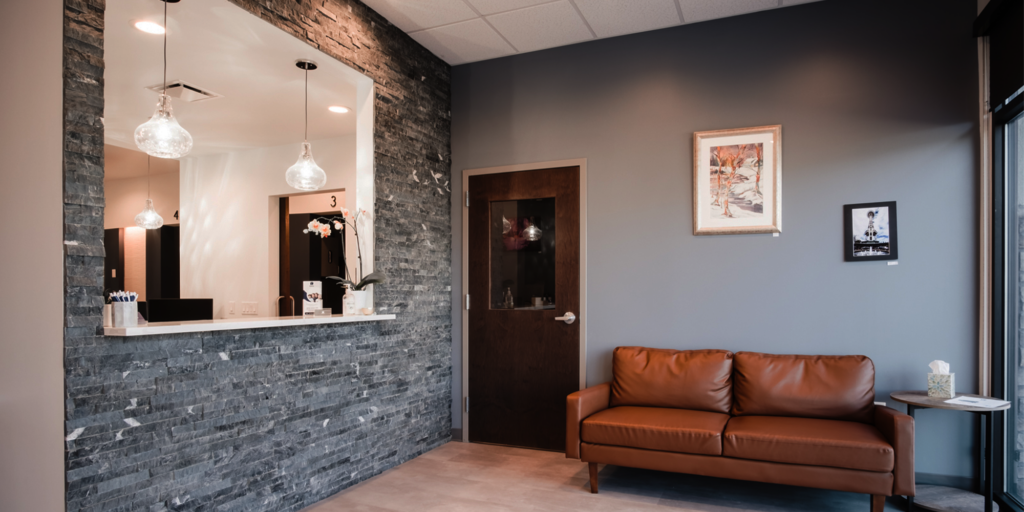 The image depicts a waiting area with a reception counter featuring a stone wall backdrop and three hanging glass light fixtures. Nearby, there is a brown leather couch, a wooden door with a glass panel, and two framed artworks decorating the walls.