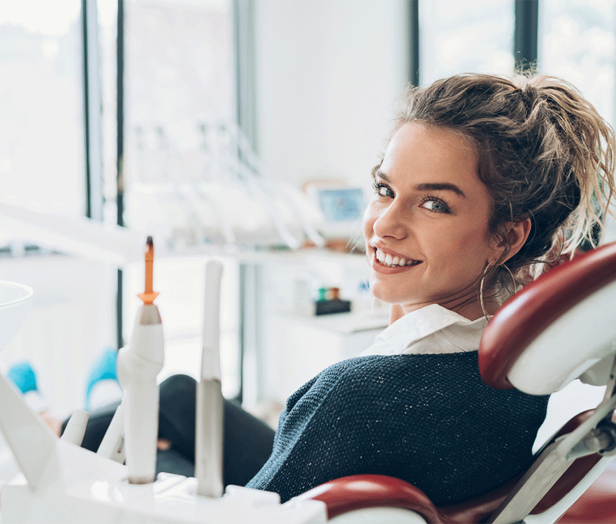 A woman sitting in a dental chair in a clinical setting. The woman's face is blurred for privacy. The background includes dental equipment and tools, indicating a dental clinic environment.