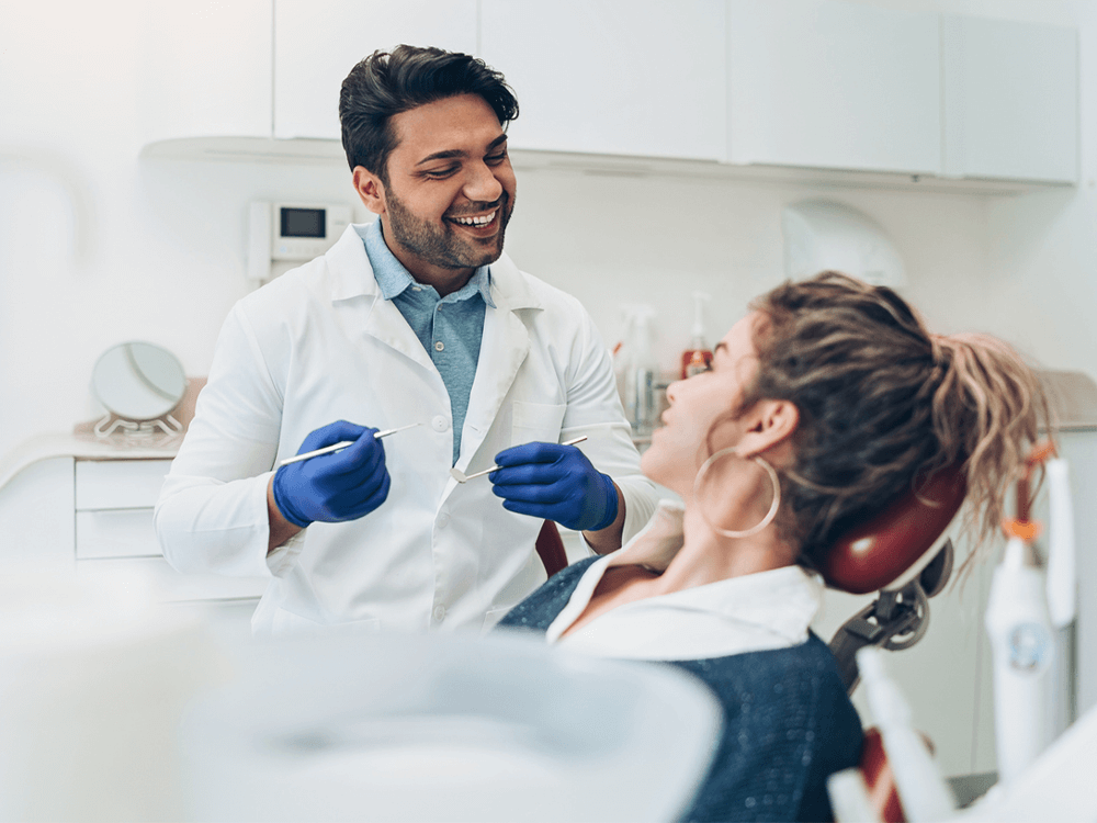 Dental clinic setting with a dentist in a white coat and blue gloves attending to a patient in a dental chair. The background includes typical dental office equipment and cabinetry.