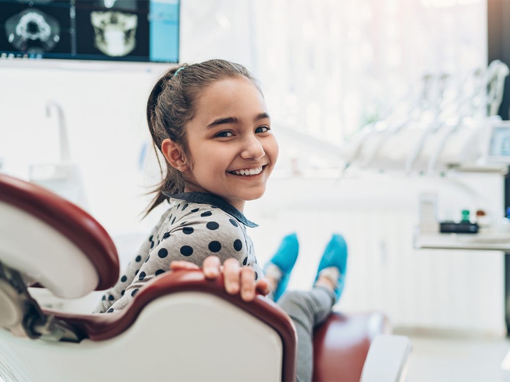 A young girl wearing a polka dot shirt and blue socks, sitting in a dental chair at a modern dental clinic. Dental instruments and a monitor displaying X-rays or scans are visible in the background.