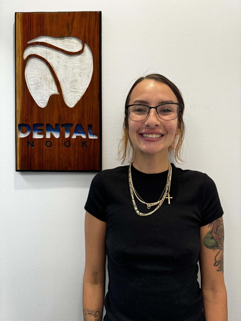 A woman standing in front of a wall with a sign that reads "DENTAL NOOK." The sign has a wooden background with a white tooth design and the text "DENTAL" in blue and "NOOK" in black. She is wearing a black shirt and multiple gold necklaces, including one with a cross pendant.